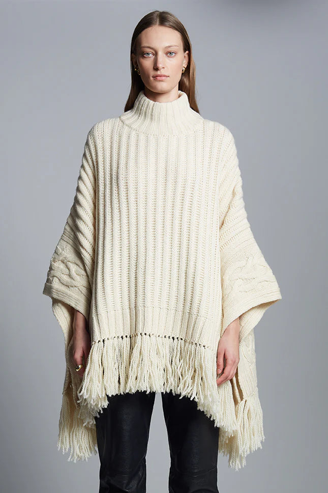 Eleven Six Hanni Poncho in 100% Peruvian wool, featuring rib and cable border stitch, fringe hem, and mock neck, shown on model layered over sweater.